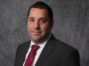 Professional headshot of a man in a dark suit with a red tie against a grey mottled background.